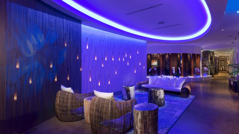 A modern lounge with ambient purple lighting, wicker chairs, and hanging lights, creating a cozy and elegant atmosphere.