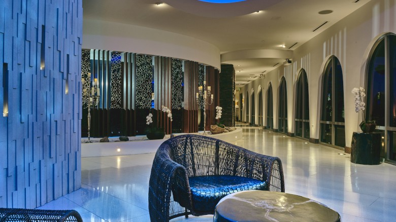 A modern, elegant lobby with curved architecture, blue lighting, wicker chairs, and decorative wall panels.