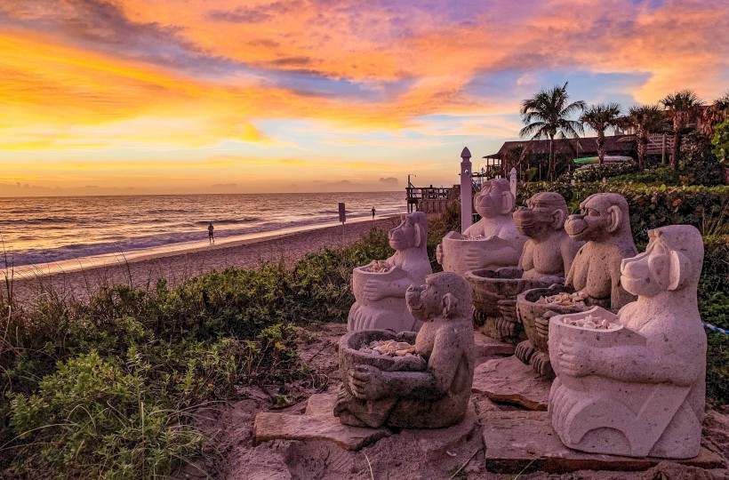 Stone sculptures sit on a sandy beach at sunset, with vibrant skies and a calm sea in the background.