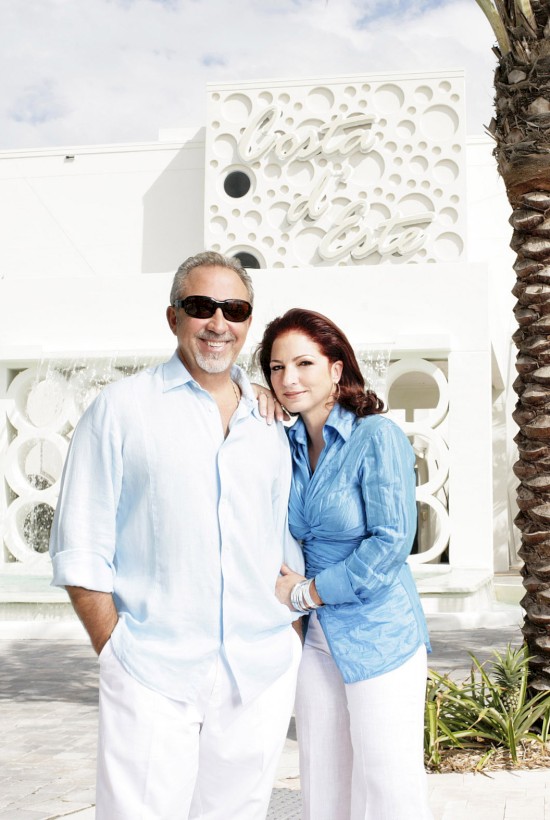 A couple in light clothing stands smiling in front of a white building and palm tree, with decorative circular patterns on the facade.