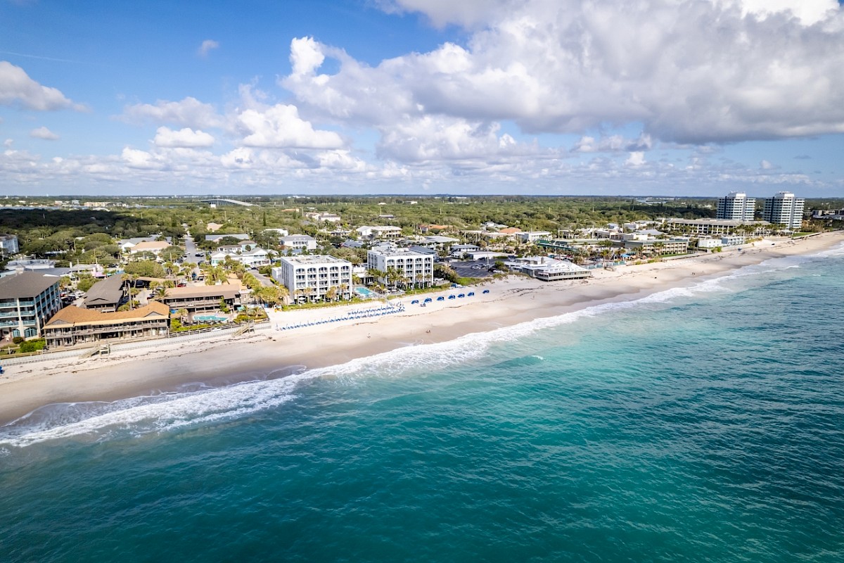 Aerial view of a coastline featuring a sandy beach, buildings, and lush greenery under a partly cloudy sky.