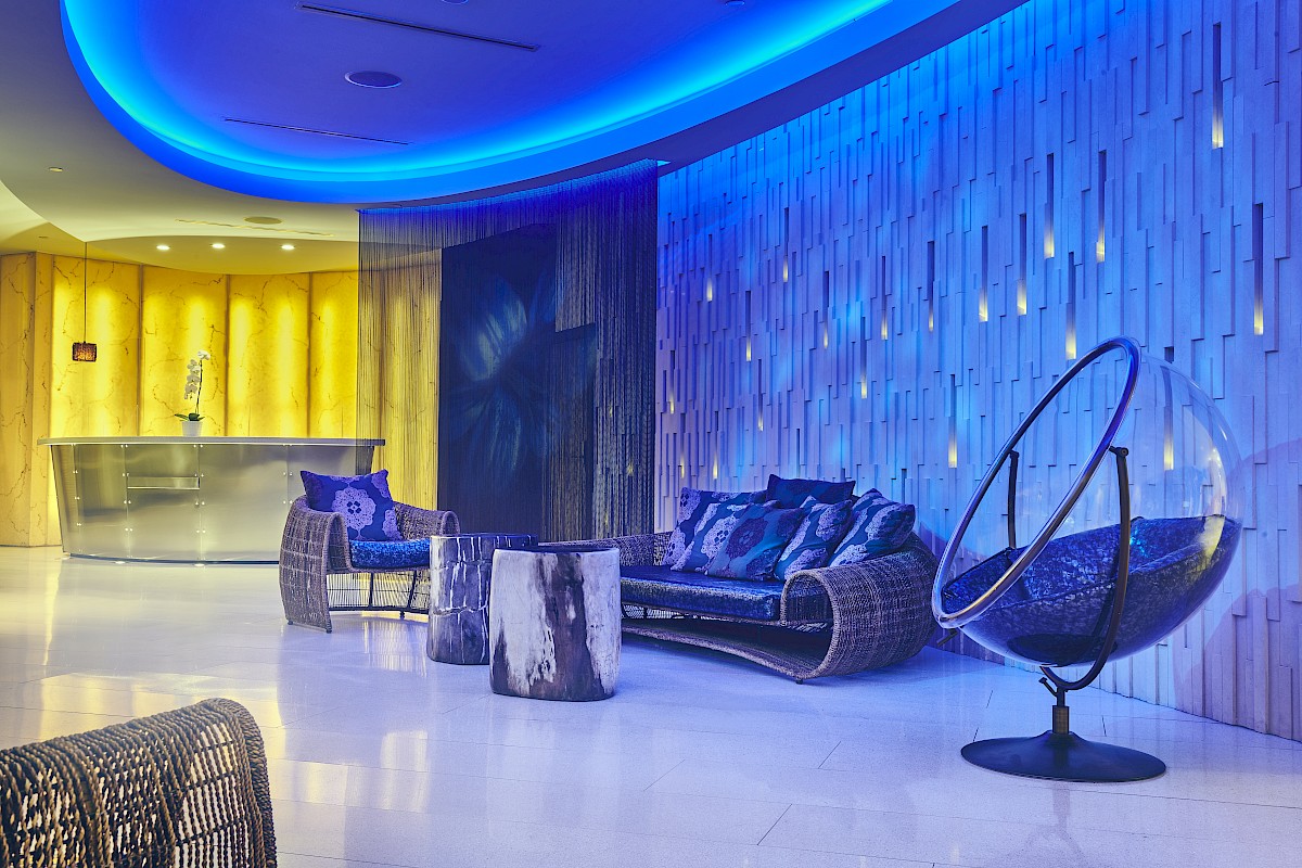 A modern lounge with blue lighting, featuring woven chairs, a sofa, a bubble chair, a tree stump table, and a sleek bar in the background.