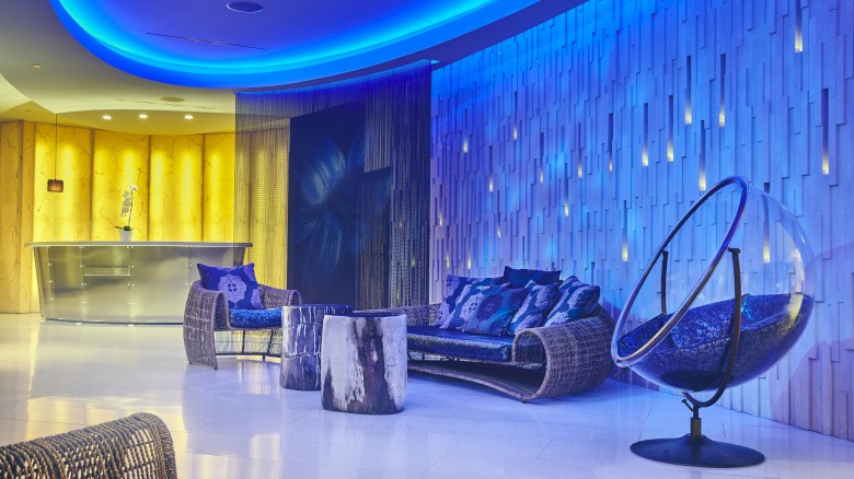 A modern lounge with blue lighting, featuring woven chairs, a sofa, a bubble chair, a tree stump table, and a sleek bar in the background.