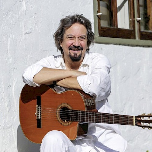 A person is smiling, sitting outside with an acoustic guitar, leaning against a white wall and near wooden-framed windows.