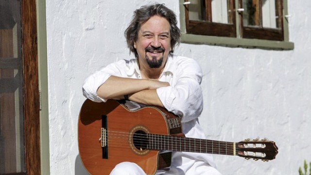 A person is smiling, sitting outside with an acoustic guitar, leaning against a white wall and near wooden-framed windows.