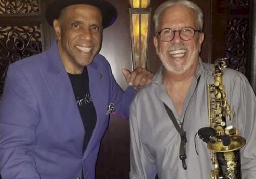 Two smiling men, one holding bongos, the other a saxophone, in a music-themed setting, wearing casual attire.