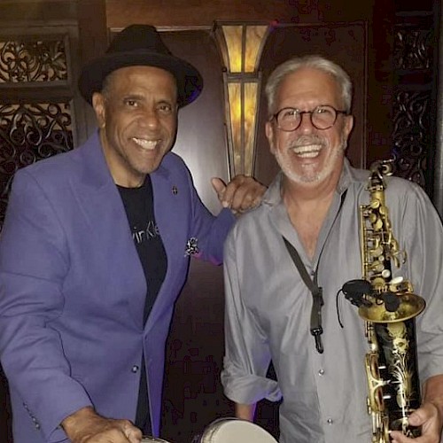 Two smiling men, one holding bongos, the other a saxophone, in a music-themed setting, wearing casual attire.