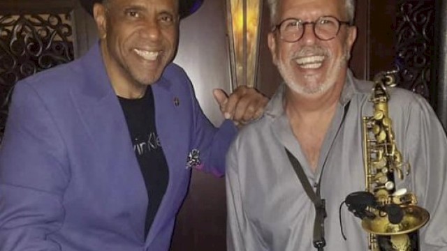 Two smiling men, one holding bongos, the other a saxophone, in a music-themed setting, wearing casual attire.