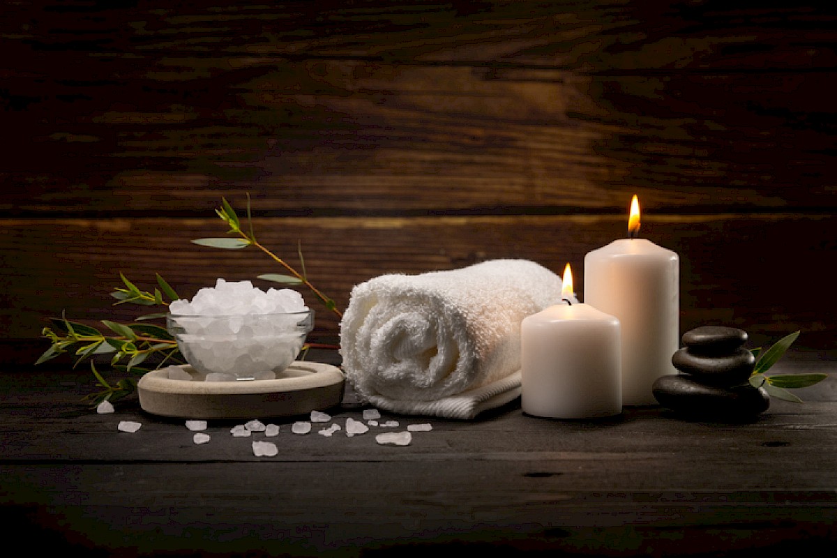 A relaxing spa scene with a rolled towel, a bowl of bath salts, scattered crystals, green sprigs, and lit candles on a dark wooden background.