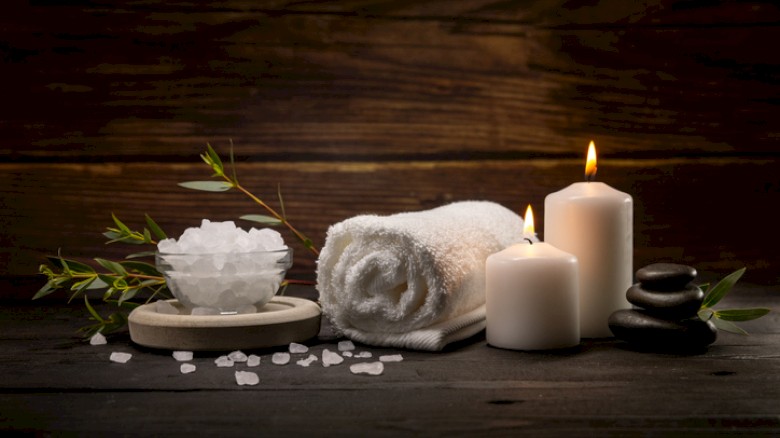 A relaxing spa scene with a rolled towel, a bowl of bath salts, scattered crystals, green sprigs, and lit candles on a dark wooden background.
