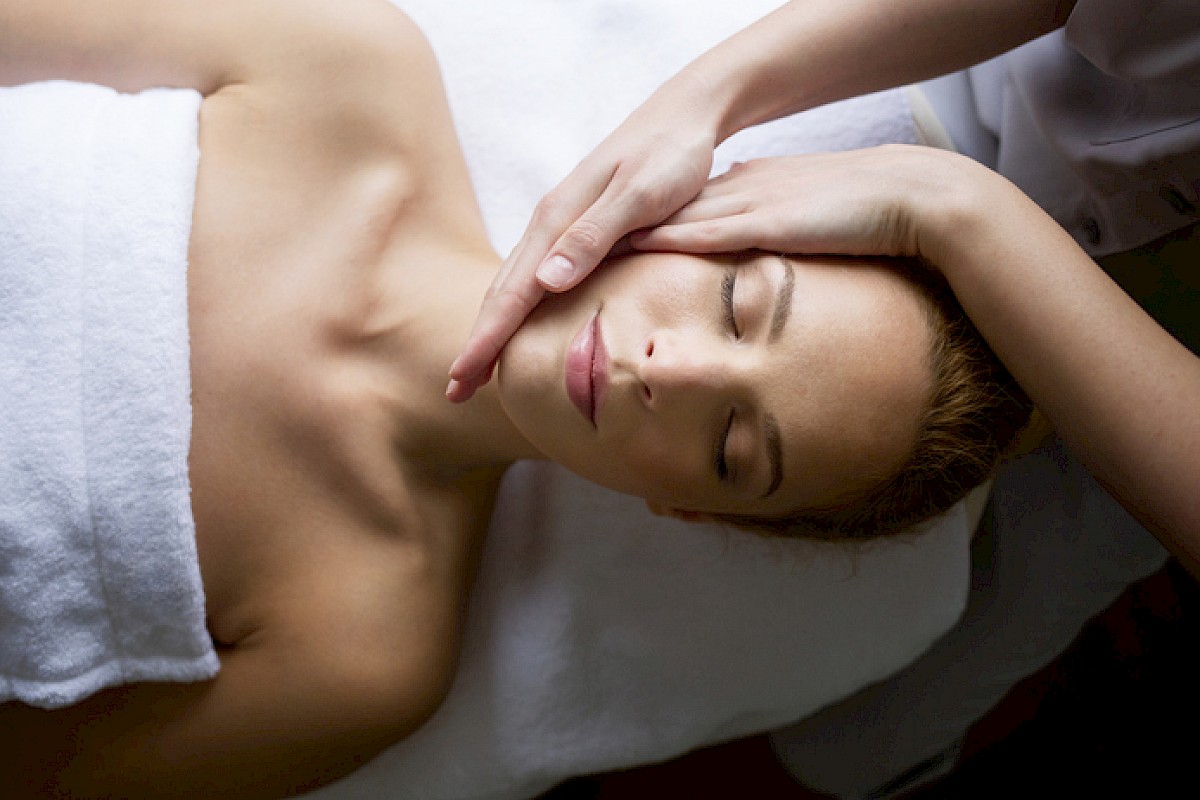 A person receives a facial/pressure massage while lying down with eyes closed, serene expression, pampering treatment in a spa context.