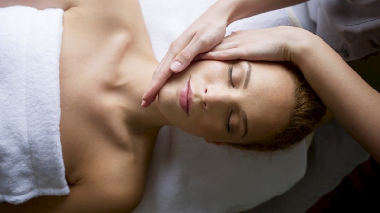 A person receives a facial/pressure massage while lying down with eyes closed, serene expression, pampering treatment in a spa context.