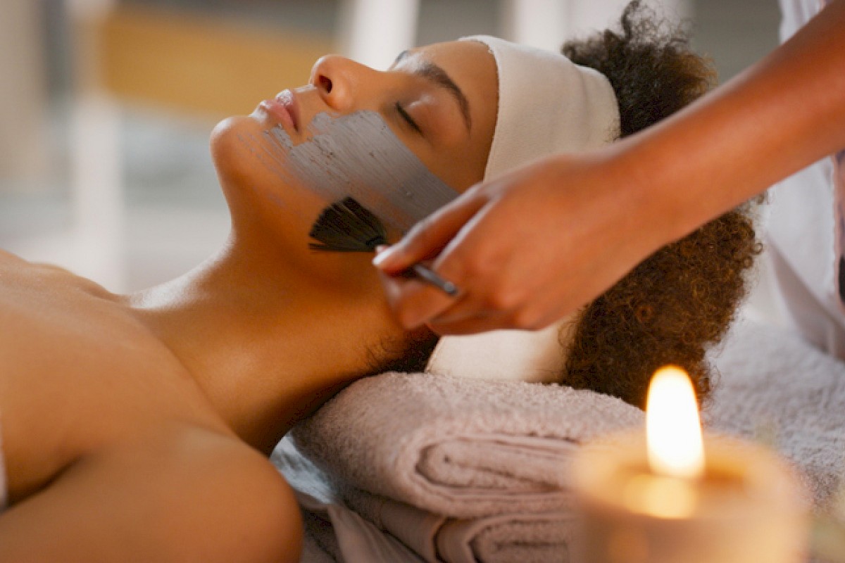 A woman is receiving a facial treatment at a spa, with a professional applying a mask or scrub on her face, near a lit candle.