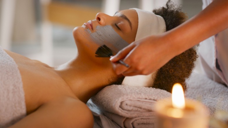A woman is receiving a facial treatment at a spa, with a professional applying a mask or scrub on her face, near a lit candle.
