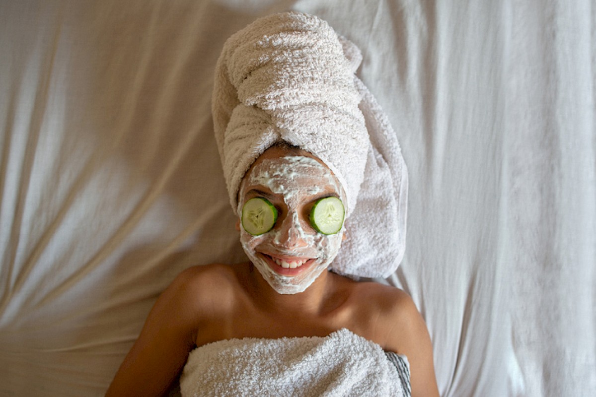 A person lying on a bed with a towel wrapped like a turban, cucumber slices over eyes, a facial mask, and a towel wrapped around the body, smiling.