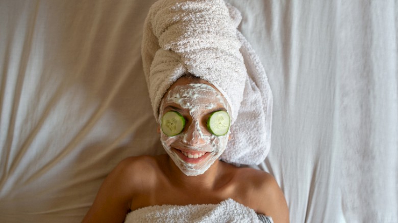 A person lying on a bed with a towel wrapped like a turban, cucumber slices over eyes, a facial mask, and a towel wrapped around the body, smiling.