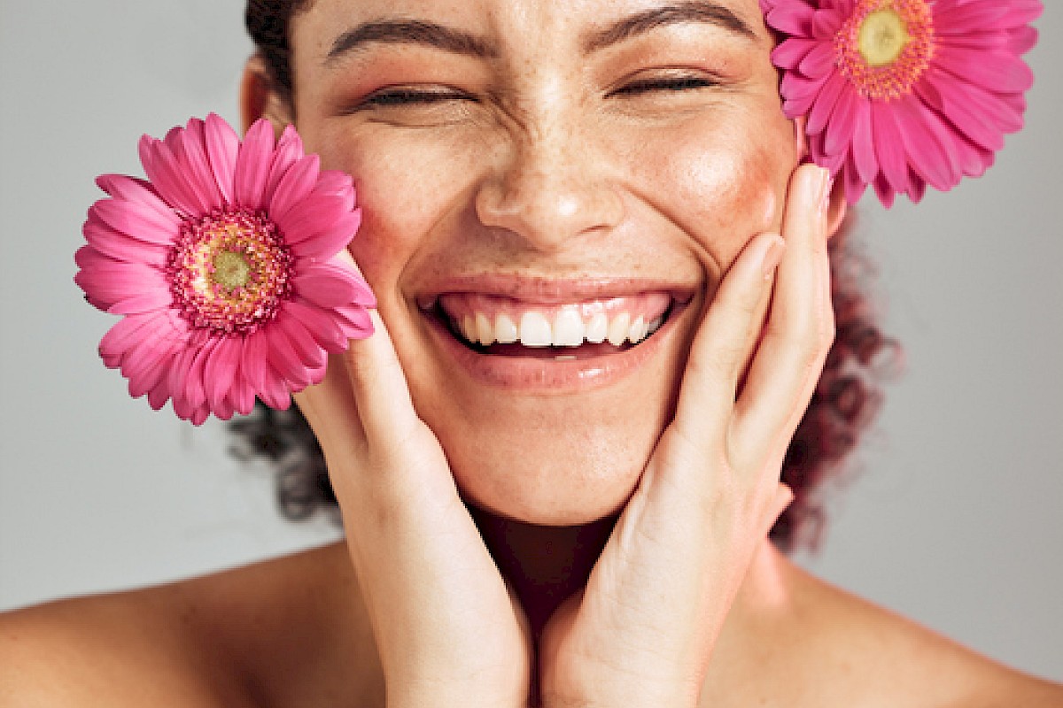 A smiling woman with pink flowers on her face, expressing happiness and joy with her eyes closed.