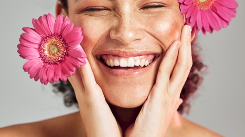 A smiling woman with pink flowers on her face, expressing happiness and joy with her eyes closed.