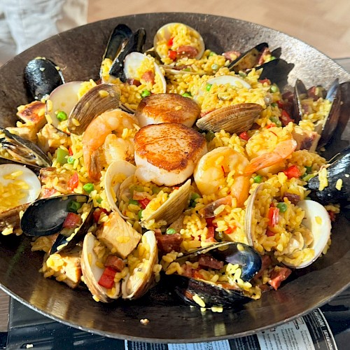 This image shows a pan filled with seafood paella, featuring mussels, clams, shrimp, scallops, and rice, garnished with peas and peppers.