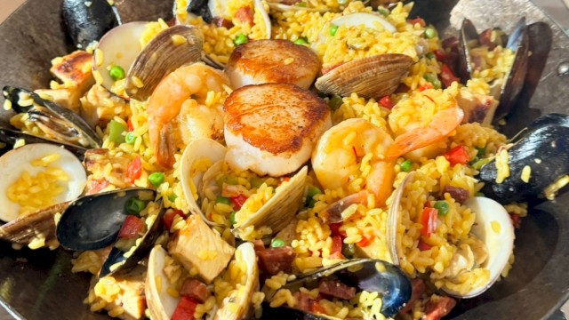 A seafood paella-style dish with mussels, clams, shrimp, scallops, and chunks of fish over saffron rice, colorful vegetables, and herbs.