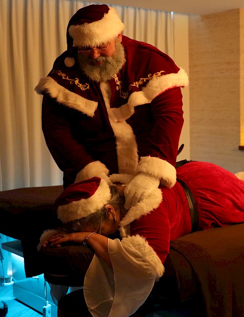 A person in a Santa suit gives a massage to another Santa-suited person in a cozy room with candles.