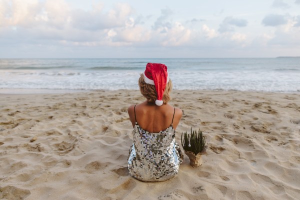 A person in a Santa hat and sequin dress sits on a sandy beach near the ocean, with a small plant beside them, facing calm waters.