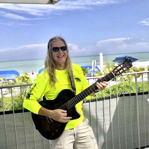 A person with long hair and sunglasses plays a guitar on a sunny beachside deck, wearing a bright yellow shirt, next to the ocean.