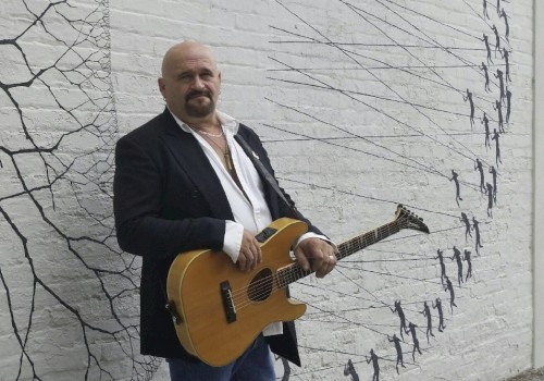 A person with a guitar stands against a mural-covered wall depicting small figures and lines, dressed in a dark jacket and jeans.