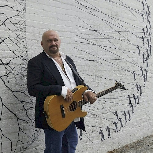 A person with a guitar stands against a mural-covered wall depicting small figures and lines, dressed in a dark jacket and jeans.