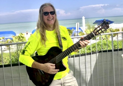 A person in sunglasses and a neon shirt plays guitar by the beach, with blue umbrellas and ocean in the background.