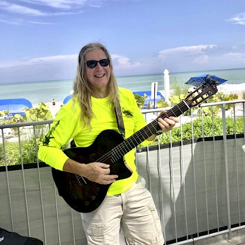 A person in sunglasses and a neon shirt plays guitar by the beach, with blue umbrellas and ocean in the background.
