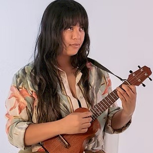 A person playing a ukulele with a thoughtful expression, wearing a floral shirt, against a plain background.