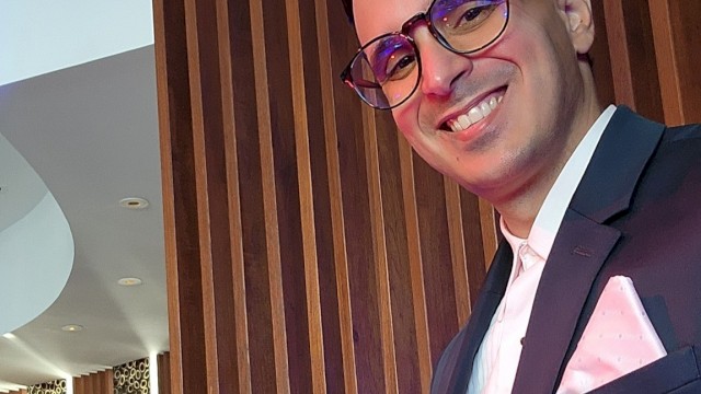 The image shows a person in a suit with glasses, smiling in a stylish setting with wood paneling and leopard print details on the walls.