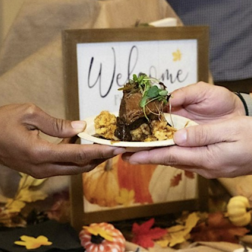 A person is receiving a piece of cake or dessert on a plate at an event, with fall-themed decorations and a welcome sign in the background.