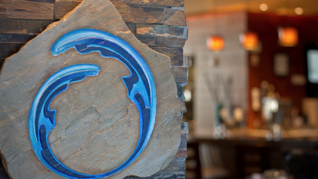 A stone plaque with a blue, circular wave design is on a wall, with a blurred interior and hanging lights in the background.