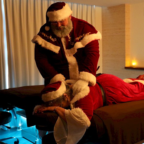 Two people dressed as Santa Claus giving or performing a massage on a table in a dimly lit room with candles and blue lighting.