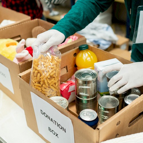A person wearing gloves is organizing donation boxes filled with canned food, pasta, juice, and toys, labeled 