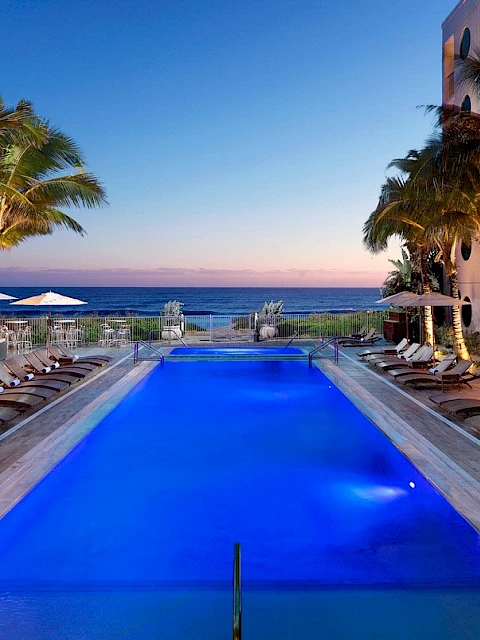 A luxurious pool lined with lounge chairs, surrounded by palm trees, overlooking the ocean at sunset, creating a serene atmosphere.