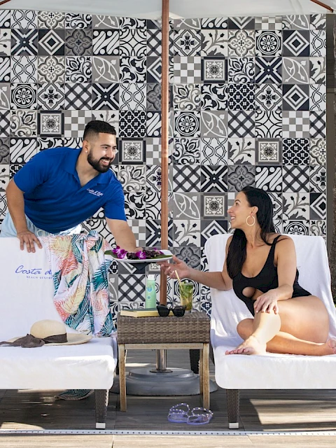 A person in a blue shirt serves a drink to a woman lounging on a sunbed, with colorful tiles in the background.