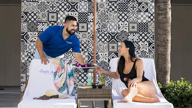 A person in a blue shirt serves a drink to a woman lounging on a sunbed, with colorful tiles in the background.