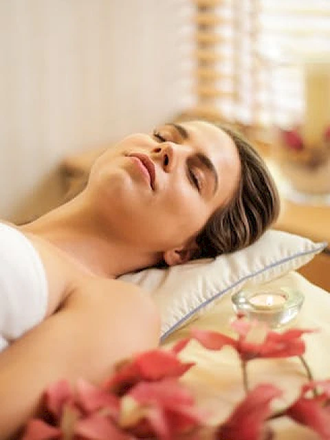 A person is relaxing on a massage table surrounded by flowers and soft lighting, suggesting a serene spa environment.