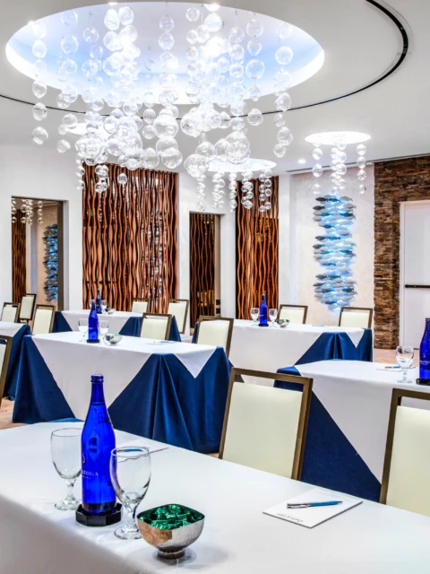 The image shows a modern conference room with draped tables, blue bottles, glasses, and stylish lighting fixtures hanging from the ceiling.