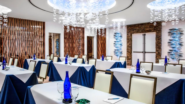 The image shows a modern conference room with draped tables, blue bottles, glasses, and stylish lighting fixtures hanging from the ceiling.