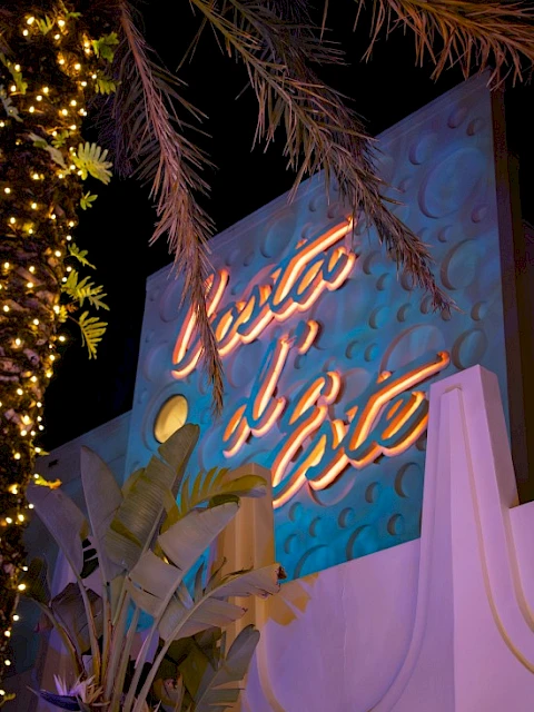 The image shows a brightly lit sign with cursive writing on a decorated wall, surrounded by palm trees and nighttime lighting.