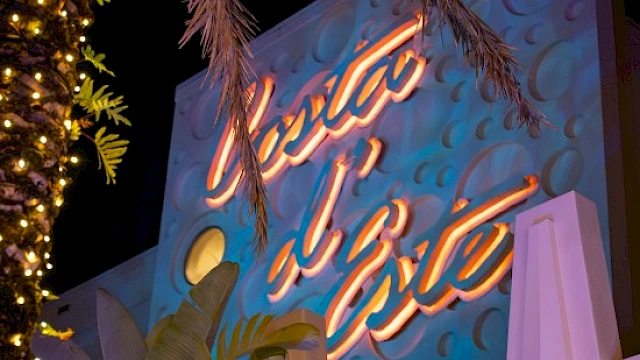 The image shows a brightly lit sign with cursive writing on a decorated wall, surrounded by palm trees and nighttime lighting.
