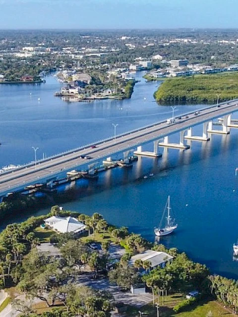 A bridge spans over a blue waterway with surrounding greenery and boats moored near the shore, leading to a distant townscape.