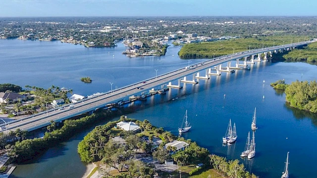A bridge spans over a blue waterway with surrounding greenery and boats moored near the shore, leading to a distant townscape.