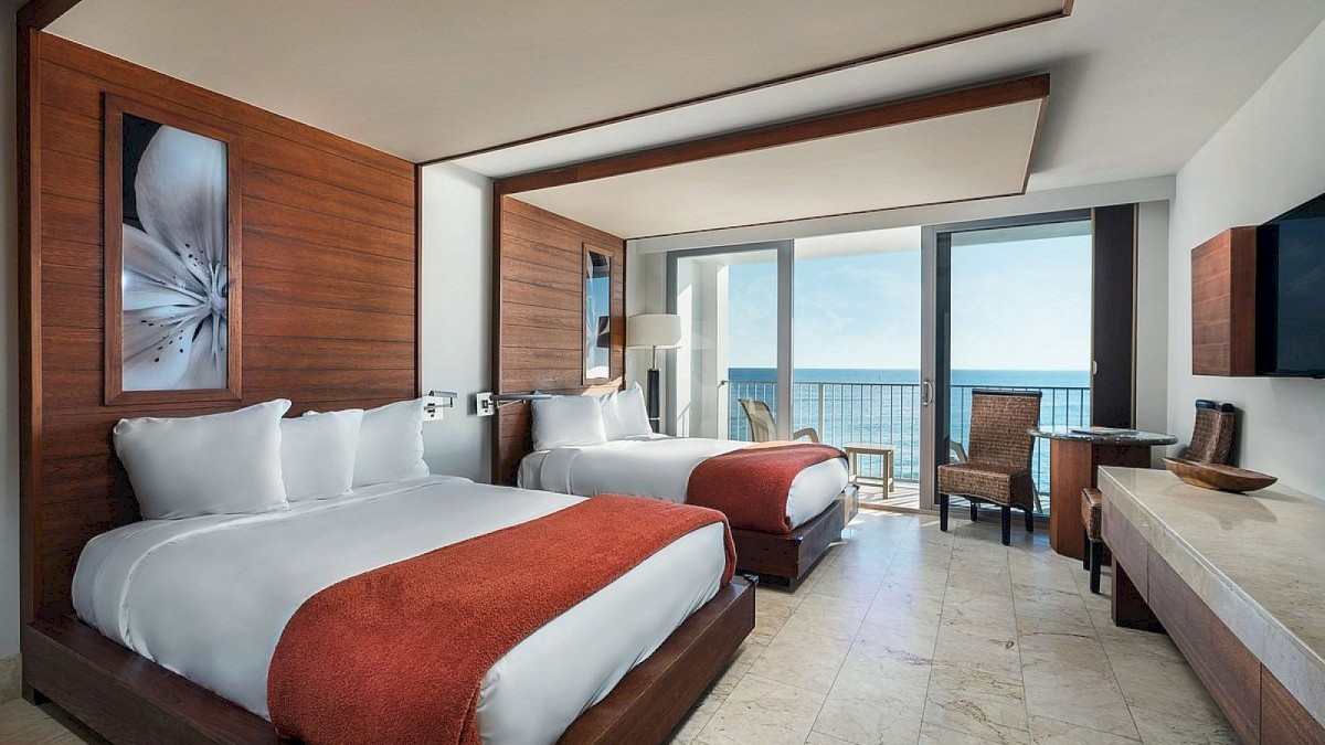 The image shows a hotel room with two beds, orange accents, a sea view, and modern furnishings, including a TV and desk, ending the sentence.