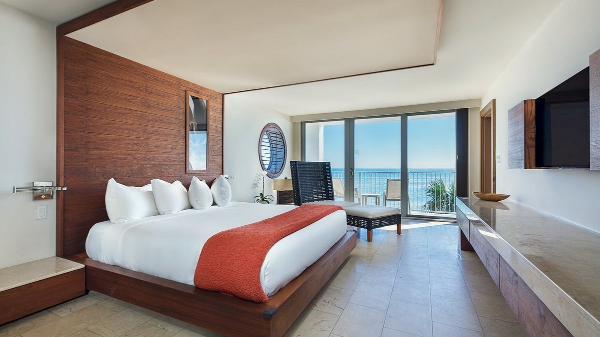 A modern hotel room features a large bed, wooden decor, a seating area, and a balcony with a view of the ocean.