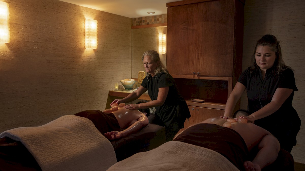 Two people receiving massages from two therapists in a dimly lit spa room with wall sconces and soft blankets on the tables.